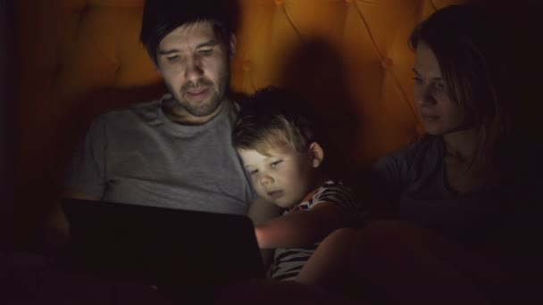 Familia feliz con su pequeño hijo aprendiendo a jugar tableta acostado en la cama en casa por la noche — Vídeos de Stock