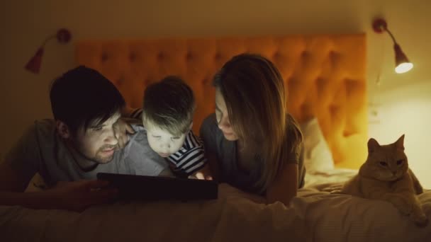 Familia feliz con su pequeño hijo y divertido gato acostado en la cama en casa y navegar por las redes sociales en la tableta antes de dormir — Vídeo de stock