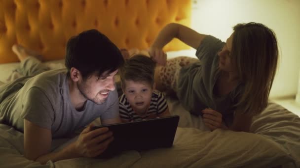 Famiglia felice con il piccolo figlio sdraiato a letto a casa e navigare sui social media sul tablet prima di dormire — Video Stock