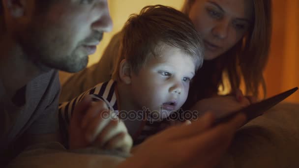 Крупный план счастливой молодой семьи, лежащей в постели дома, в то время как маленький мальчик учится играть в цифровой планшетный компьютер перед сном — стоковое видео