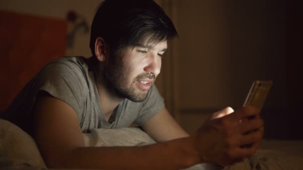 Närbild av Young leende man använder smartphone för surfing sociala media ligger i sängen hemma på natten — Stockvideo