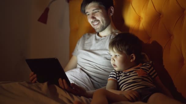 Junger Vater und sein kleiner Sohn sehen Zeichentrickfilm mit Tablet-Computer, während sie abends zu Hause im Bett liegen — Stockvideo