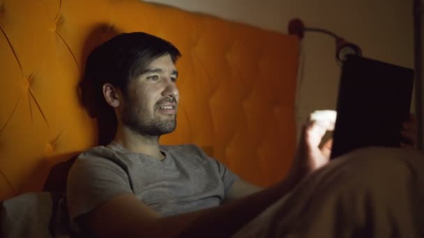 Młody człowiek uśmiechający się przy użyciu komputera typu tablet dla mediów społecznych surfing, leżąc w łóżku w domu przed snem — Wideo stockowe