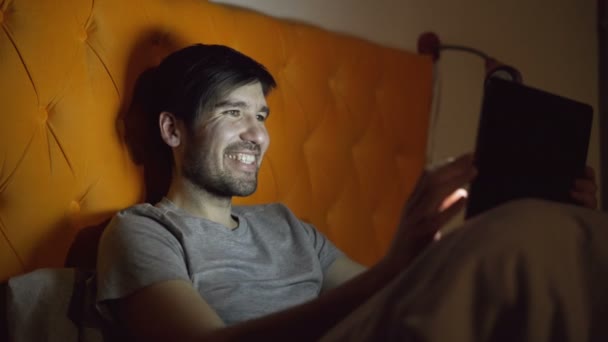 寝る前に自宅のベッドで横になっているサーフィン ソーシャル メディアのタブレット コンピューターを使用して若い笑い男 — ストック動画