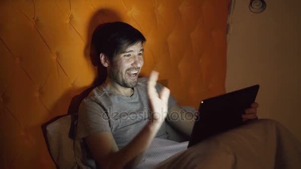 寝る前に自宅のベッドで横になっている中にガール フレンドとオンライン ビデオ チャットを持つタブレット コンピューターを使用して陽気な若者 — ストック動画