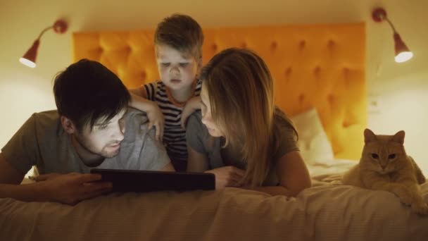 Familia feliz con su pequeño hijo y divertido gato acostado en la cama en casa y el uso de la tableta para jugar antes de dormir — Vídeos de Stock