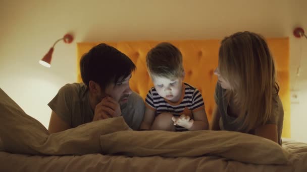 夜に自宅のベッドで横になっているタブレット コンピューターを再生する学習の幼い息子と幸せな家庭 — ストック動画