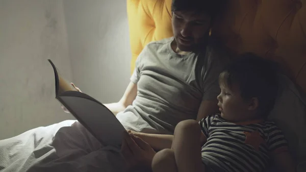 Jeune père avec un petit fils couché sur le lit à la maison et lisant un livre de contes de fées avant de dormir le soir — Photo