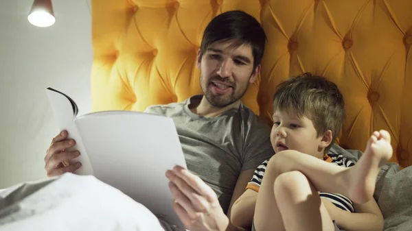 Jeune père avec un petit fils couché sur le lit à la maison et lisant un livre de contes de fées avant de dormir le soir — Photo