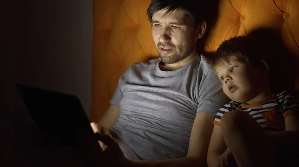 Jeune père et son petit fils regardant un film de dessin animé à l'aide d'un ordinateur tablette et parlant tout en étant couché dans le lit le soir à la maison — Photo