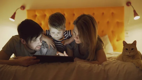 Счастливая семья с маленьким сыном и веселой кошкой, лежащей дома в постели и использующей планшетный компьютер для игры перед сном — стоковое фото