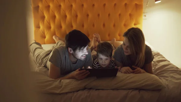 A kis fia a boldog család egy ágyban fekve, otthon, és használja a táblaszámítógép néz film rajzfilm, lefekvés előtt — Stock Fotó