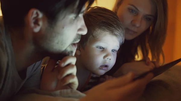 Közelkép a boldog fiatal család feküdt az ágyban otthon, míg a tanulás játék digitális tabletta számítógép idő lefekvés előtt kisfiú — Stock Fotó