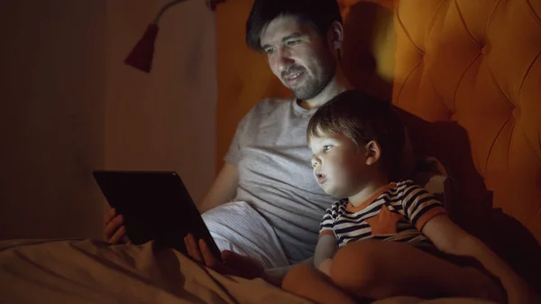 若い父親と夜の自宅でベッドに横たわっている間タブレット コンピューターを使用して漫画映画を見て彼の幼い息子 — ストック写真
