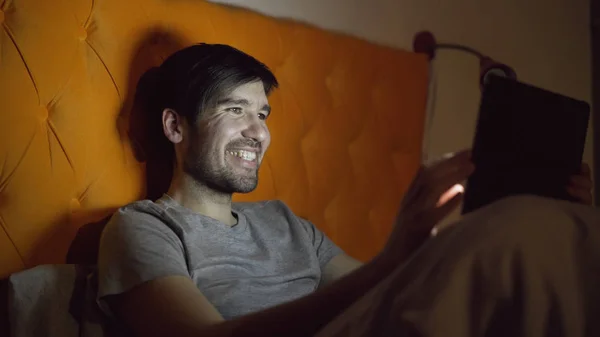 Молодой смеющийся человек, пользующийся планшетным компьютером для серфинга в социальных сетях, лежащих в постели перед сном — стоковое фото