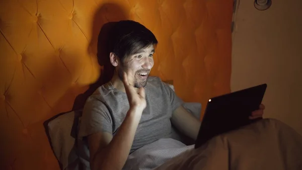 Jovem homem alegre usando computador tablet ter vídeo chat on-line com a namorada enquanto deitado na cama em casa antes de dormir — Fotografia de Stock