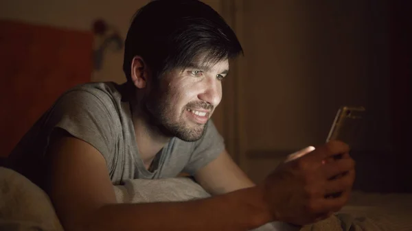 Close-up de Jovem sorrindo homem usando smartphone para navegar nas redes sociais deitado na cama em casa à noite — Fotografia de Stock