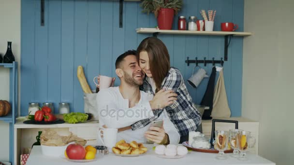 Casal atraente se encontram na cozinha na hora do café da manhã e usando smartphone para surfar em casa — Vídeo de Stock