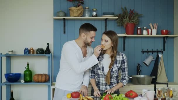 Feliz pareja joven en la cocina. Atractivo hombre bailando alimentando a su novia mientras ella está cocinando por la mañana — Vídeo de stock