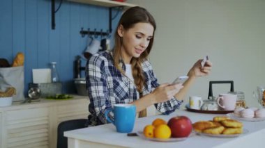 Gülümseyen kadın kullanarak online alışveriş yaparken smartphone ve kredi kartı süre evde mutfakta kahvaltı var