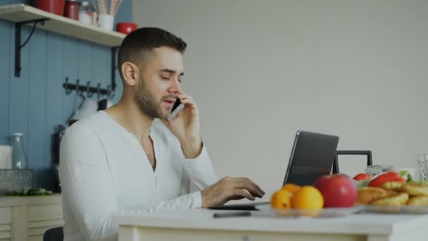 Hombre joven guapo hablando de teléfono y el uso de ordenador portátil sentado en la cocina después del desayuno por la mañana — Vídeo de stock
