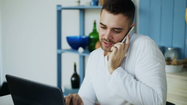 Knappe jongeman telefoon praten en het gebruik van laptopcomputer zitten in de keuken na het ontbijt in de ochtend — Stockvideo