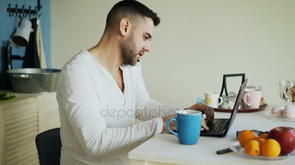 Красивый молодой человек разговаривает по телефону и с помощью ноутбука, сидя на кухне после завтрака утром — стоковое видео