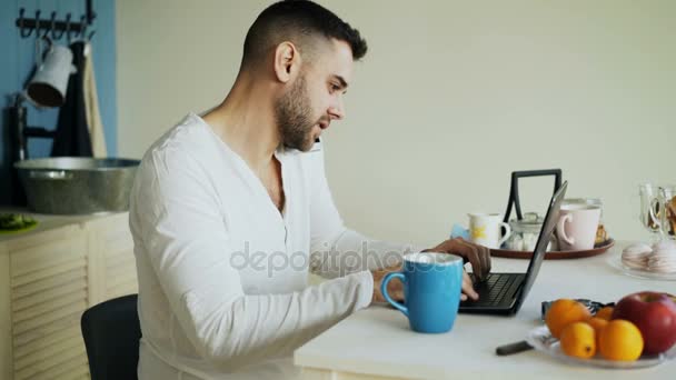 Handsome young man talking phone and using laptop computer sitting in the kitchen after breakfast in the morning — Stock Video