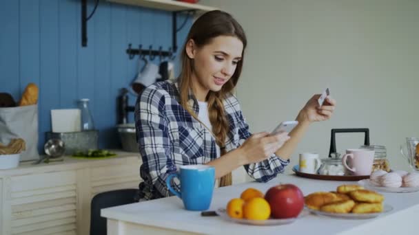 Lachende vrouw doen online winkelen via smartphone en credit card terwijl ontbijten in de keuken thuis — Stockvideo