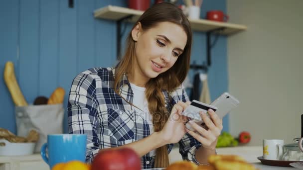 Gladlynt kvinna betala räkningar online med smartphone och kreditkort samtidigt äta frukost i köket hemma — Stockvideo