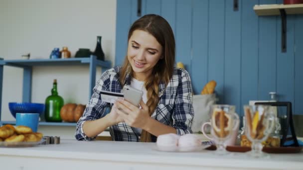 Smiling woman doing online shopping using smartphone and credit card while have breakfast in the kitchen at home — Stock Video