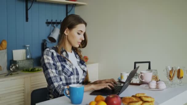 Молодая сконцентрированная женщина, работающая на кухне с ноутбуком во время завтрака по утрам — стоковое видео