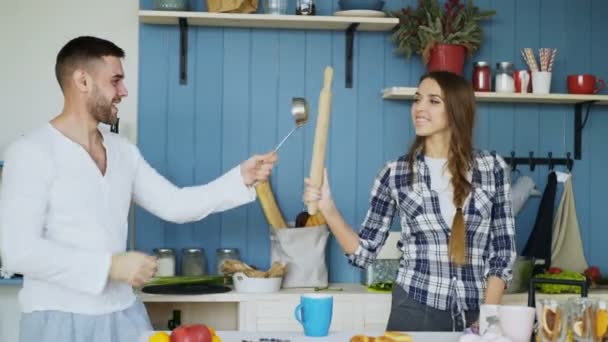 Pareja feliz divirtiéndose en la esgrima de la cocina con cucharón y rodillo mientras cocina el desayuno en casa — Vídeos de Stock