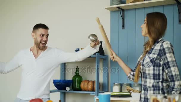 Happy couple having fun in the kitchen fencing with ladle and rolling-pin while cooking breakfast at home — Stock Video