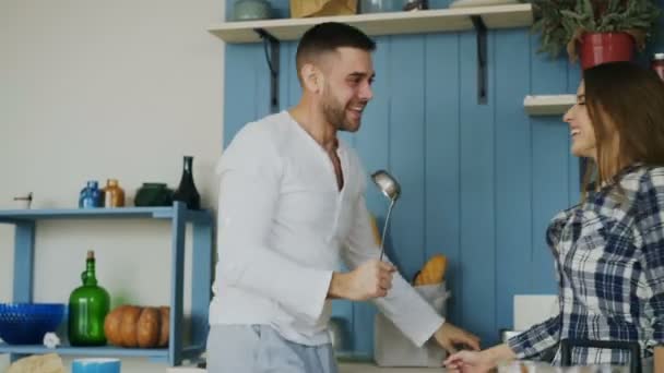 Feliz pareja joven se divierten bailando y cantando en la cocina mientras escuchan música en la mañana en casa — Vídeo de stock