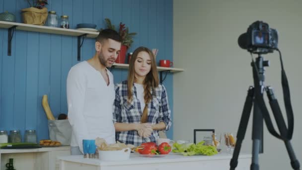 Alegre pareja atractiva grabación video blog sobre comida vegetariana saludable en la cámara dslr en la cocina en casa — Vídeo de stock