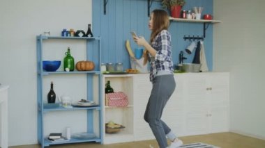 Neşeli mutlu kız dans ve sörf sosyal medya onun smartphone cep telefonu ile evde süre mutfakta sabah şarkı