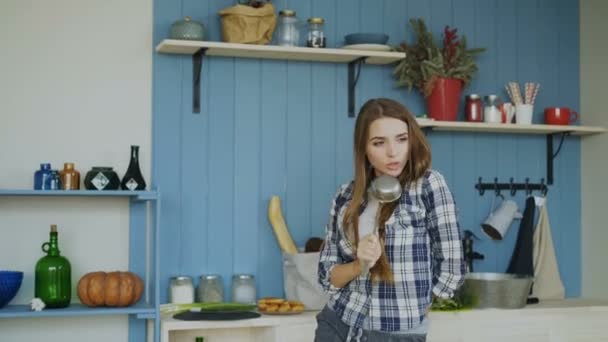 Веселая юная забавная женщина танцует и поет с ковшами во время досуга на кухне дома — стоковое видео