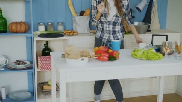 По утрам веселая девочка танцует и поет на кухне, готовя завтрак дома — стоковое видео
