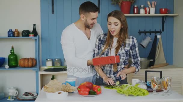 Happy cheerful man surprising his girlfriend with a gift at home in the kitchen while she cooking breakfast — Stock Video