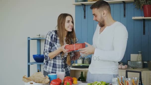 Cheerful woman surprising his boylfriend with birthday gift at home in the kitchen while he cooking breakfast — Stock Video