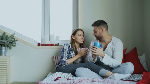 Gelukkige jonge paar praten en lachen zittend op bed en drinken koffie in de ochtend thuis — Stockvideo