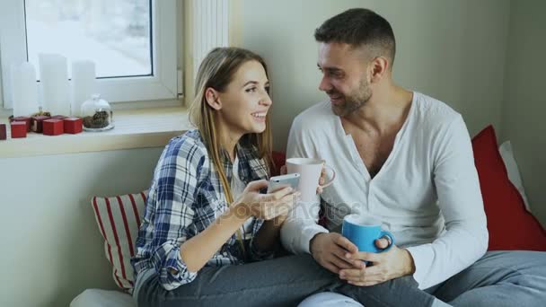 幸せな若いカップルの話し、ベッドや飲み物のコーヒーに朝は自宅座ってスマート フォンでソーシャル メディアを閲覧 — ストック動画