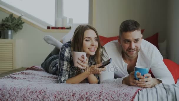 Улыбающаяся пара смотрит телевизор, лежа в постели, и пьет кофе по утрам дома — стоковое видео