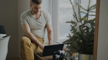 Kamera ve dizüstü bilgisayarla evde pencere kenarında oturan ve pay onun fotoğraf sosyal medya için konsantre erkek fotoğrafçı