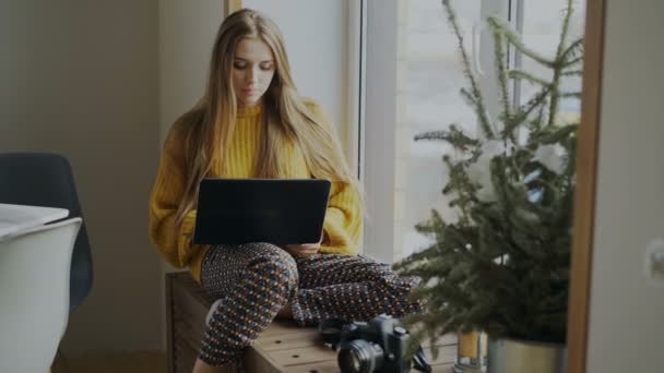 Skoncentrowany kobieta fotograf z laptopa i kamery komputera siedząc na parapecie w domu i jej udostępnianie w serwisach społecznościowych — Wideo stockowe