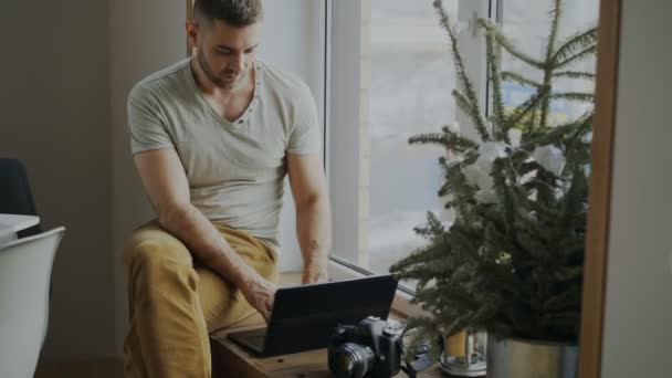 カメラとラップトップのコンピューターと自宅の窓辺に座っているソーシャル メディアに彼女の写真を共有する集中の男性カメラマン — ストック動画