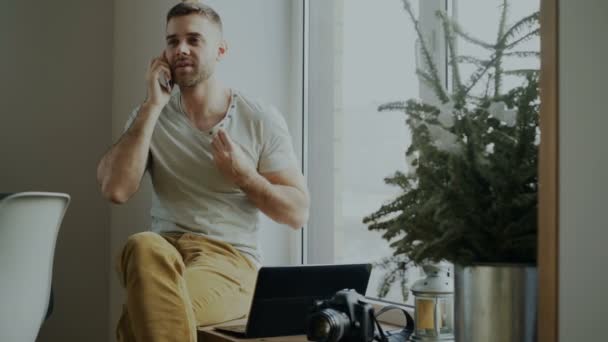 Atractivo joven hablando de teléfono sentado en el alféizar de la ventana con el ordenador portátil y la cámara en casa — Vídeo de stock