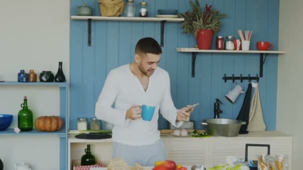 Slowmotion de joven guapo divertido bailando y cantando en la cocina mientras navega por las redes sociales en su teléfono inteligente en casa por la mañana — Vídeo de stock