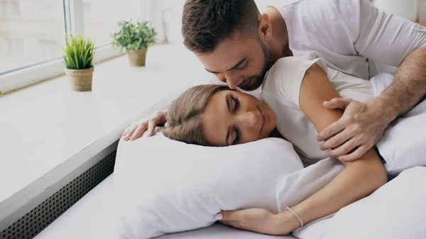 Young beautiful and loving couple wake up at the morning. Attractive man kiss and hug his wife in bed — Stock Photo, Image